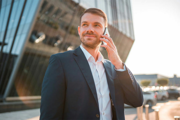 Das Bild zeigt einen Verkaufsberater im Außendienst. Er trägt Hemd und Anzug und telefoniert mit seinem Smartphone am Ohr. Im Hintergrund ist ein Gebäude und daneben einige Fahrzeuge.