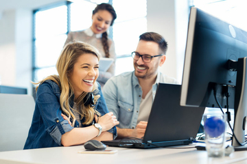 Das Bild zeigt mehrere lächelnde Personen, die am Schreibtisch sitzen und etwas am Computer ansehen.